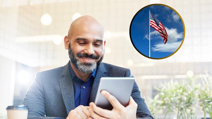 Hombre observando su dispositivo móvil en un entorno laboral y de fondo una bandera de Estados Unidos (Fotos vía Getty Images)