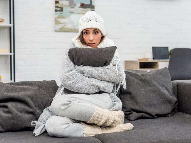 Mujer con frío, vistiendo gorro abrigo y abrazando un cojín debido a las najas temperaturas en su apartamento (Foto vía Getty Images)