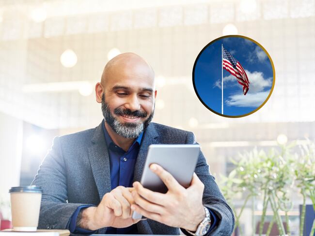 Hombre observando su dispositivo móvil en un entorno laboral y de fondo una bandera de Estados Unidos (Fotos vía Getty Images)