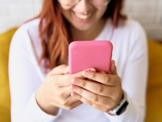 Mujer de camisa blanca utilizando aplicaciones en su celular mientras sonríe (Foto vía Getty Images)