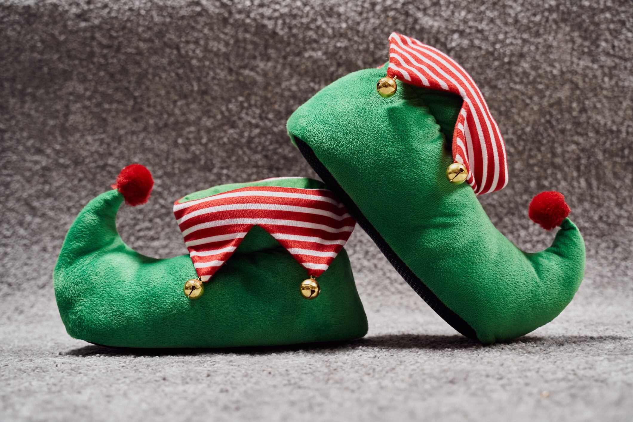 Front view of two Christmas elf novelty green slippers with bells on the rug of the living room. The green shoes with red and white stripes and a red ball at the top of the horn. Horizontal photography