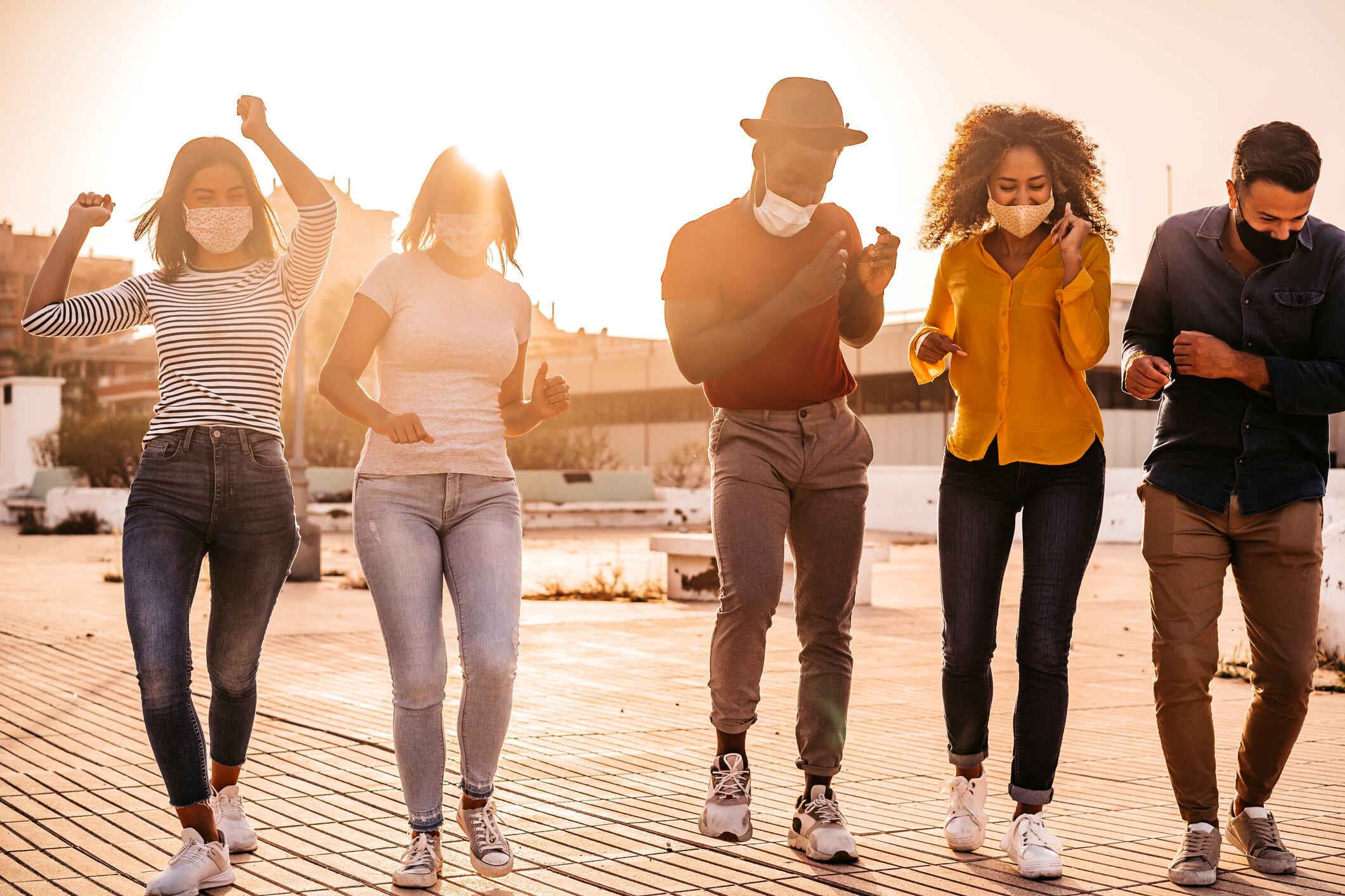 Group of young men and women, friends, wearing protective face masks and dancing at sunset.