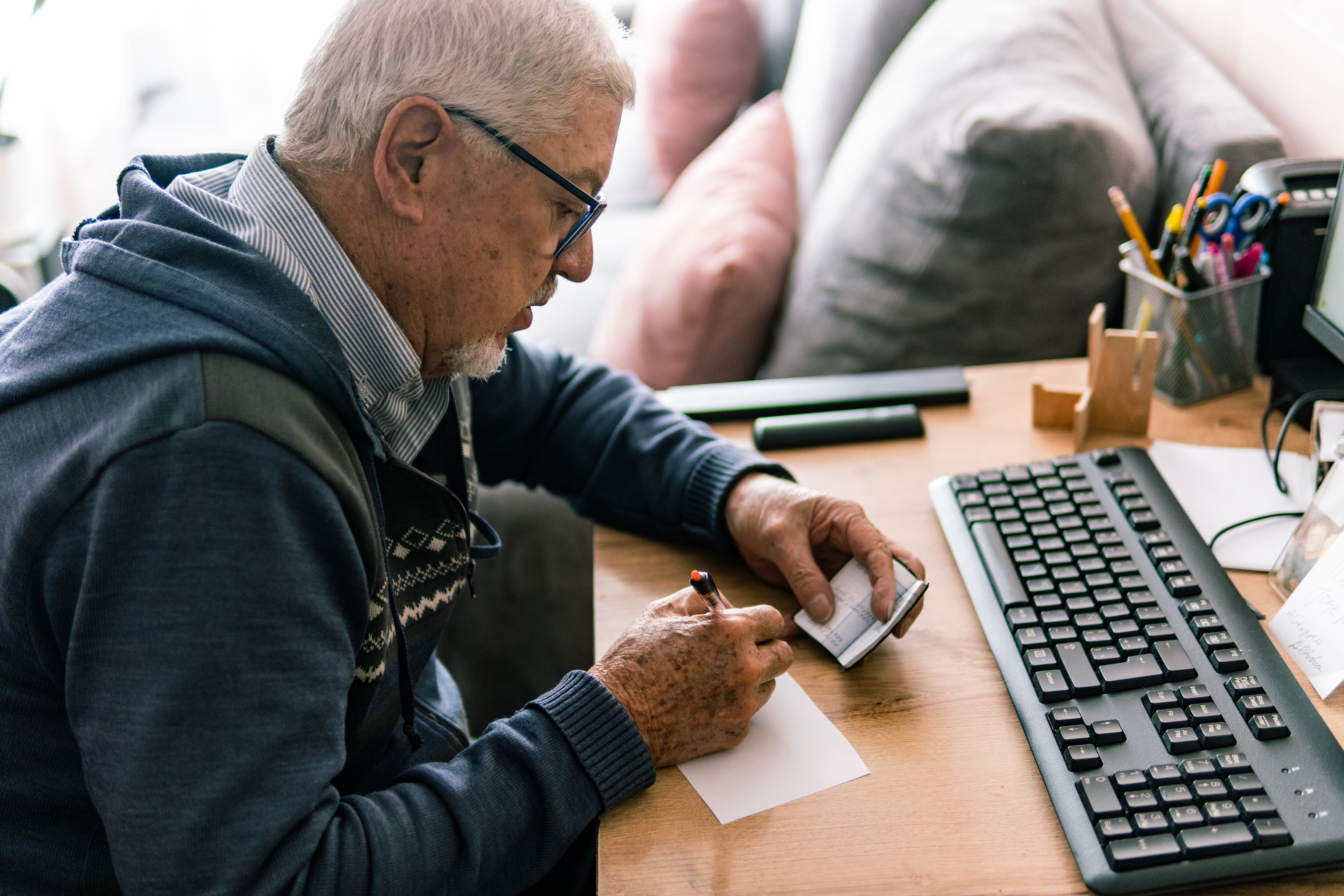 Anciano con gafas sentado en su escritorio, escribiendo en un cuaderno, con un teclado y un ordenador cerca. Getty Images