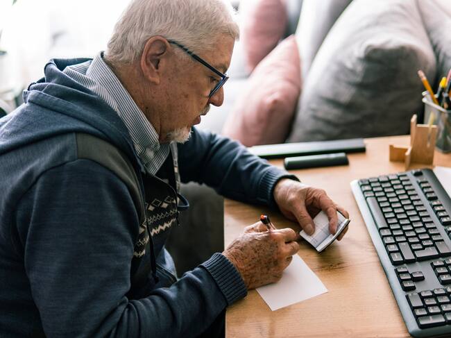 Anciano con gafas sentado en su escritorio, escribiendo en un cuaderno, con un teclado y un ordenador cerca. Getty Images