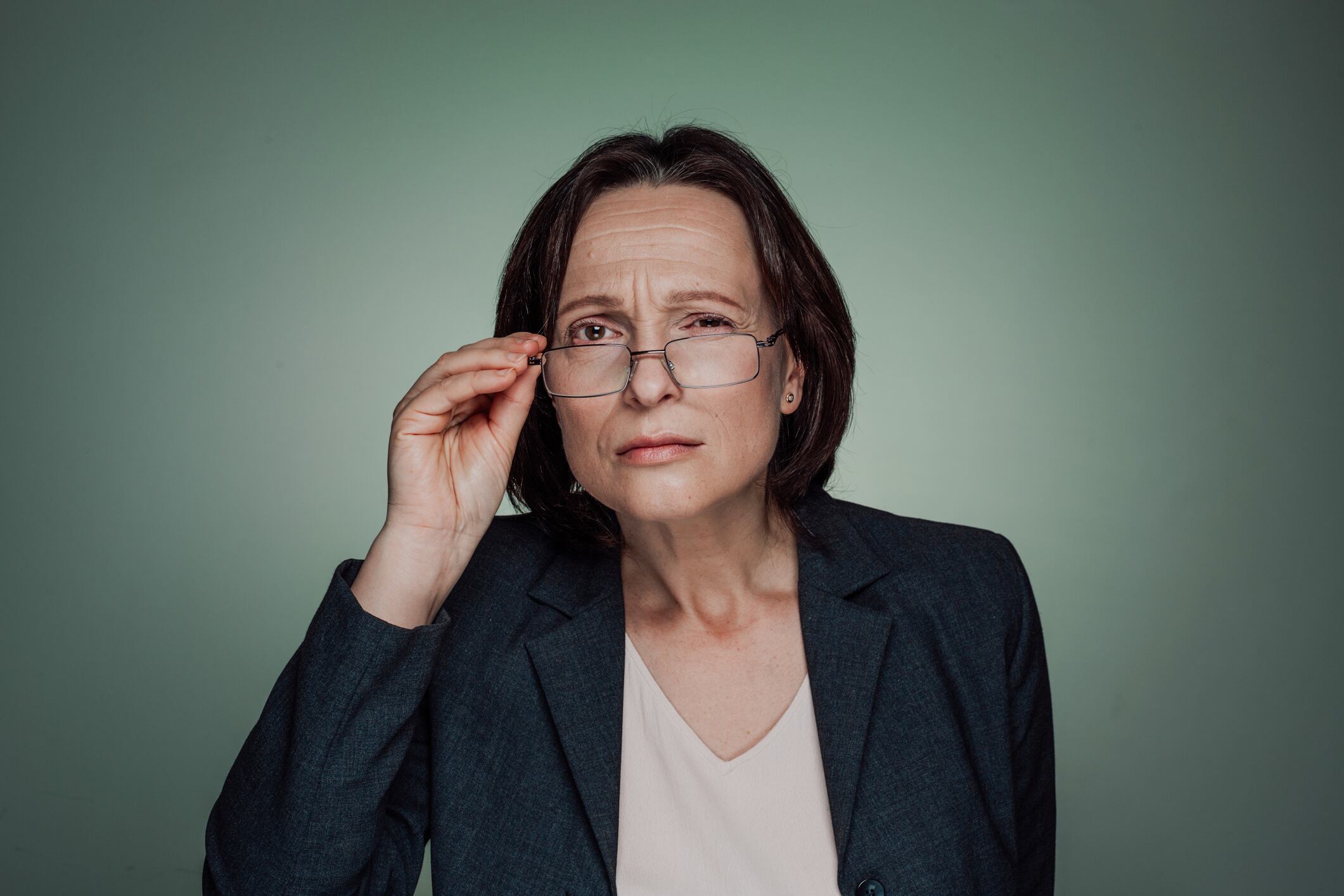 Portrait of sceptic mature adult businesswoman holding eyeglasses and looking at the camera against dark green background.