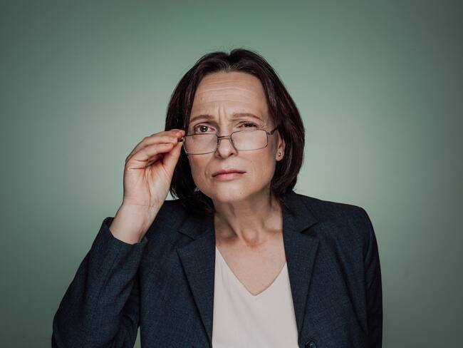 Portrait of sceptic mature adult businesswoman holding eyeglasses and looking at the camera against dark green background.