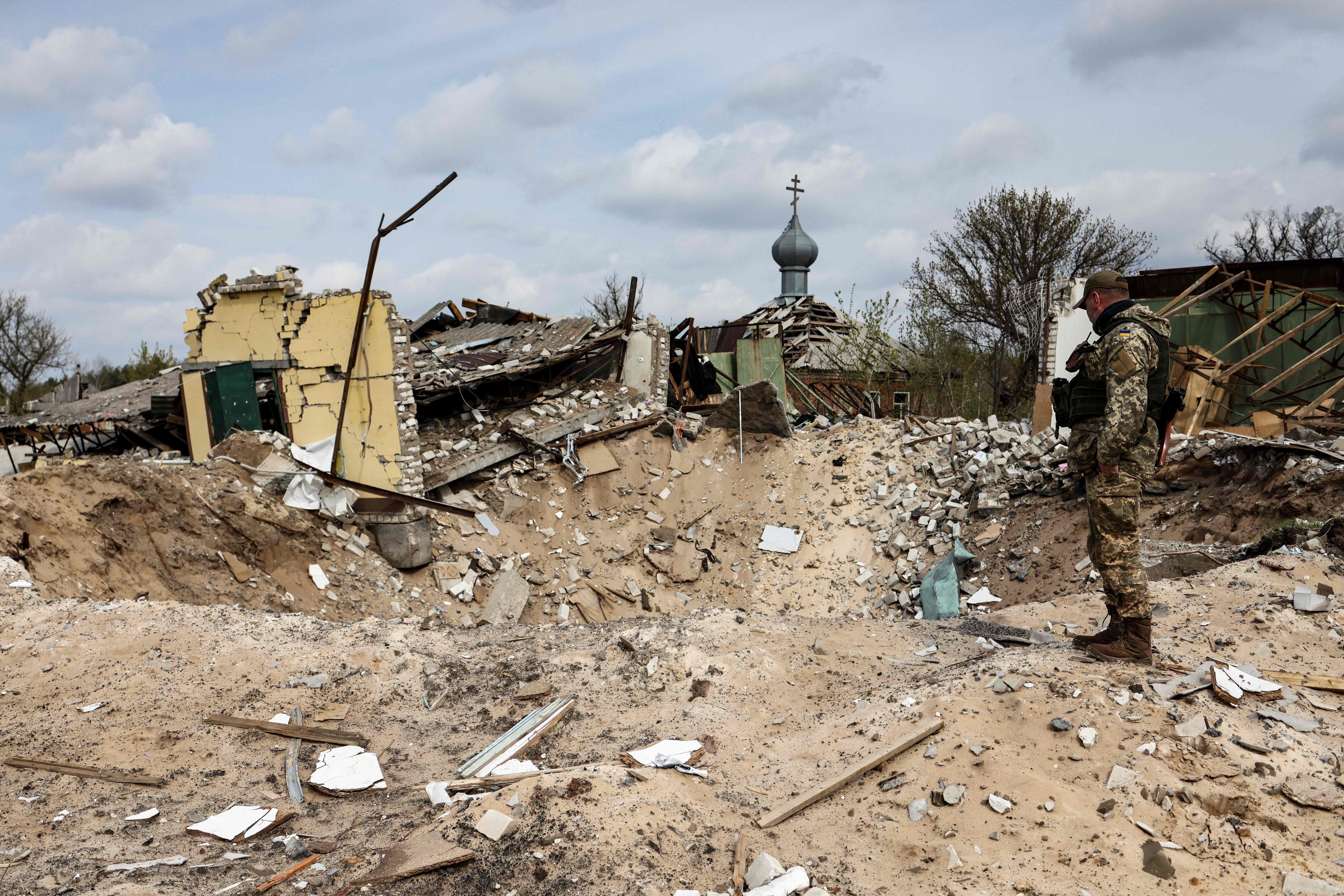 A Ukranian serviceman looks into a crater and a destroyed home are pictured in the village of Yatskivka, eastern Ukraine on April 16, 2022. - Russia's military focus now seems to be on seizing the eastern Donbas region, where Russian-backed separatists control the Donetsk and Lugansk areas. Lugansk governor Serhiy Gaidai called on April 16, 2022, for civilians to leave the area while they still can. (Photo by RONALDO SCHEMIDT / AFP) (Photo by RONALDO SCHEMIDT/AFP via Getty Images)