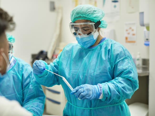 Over the shoulder view of healthcare worker in protective clothing, eyewear, bouffant cap, surgical mask, and gloves placing swab specimen in sterile container.