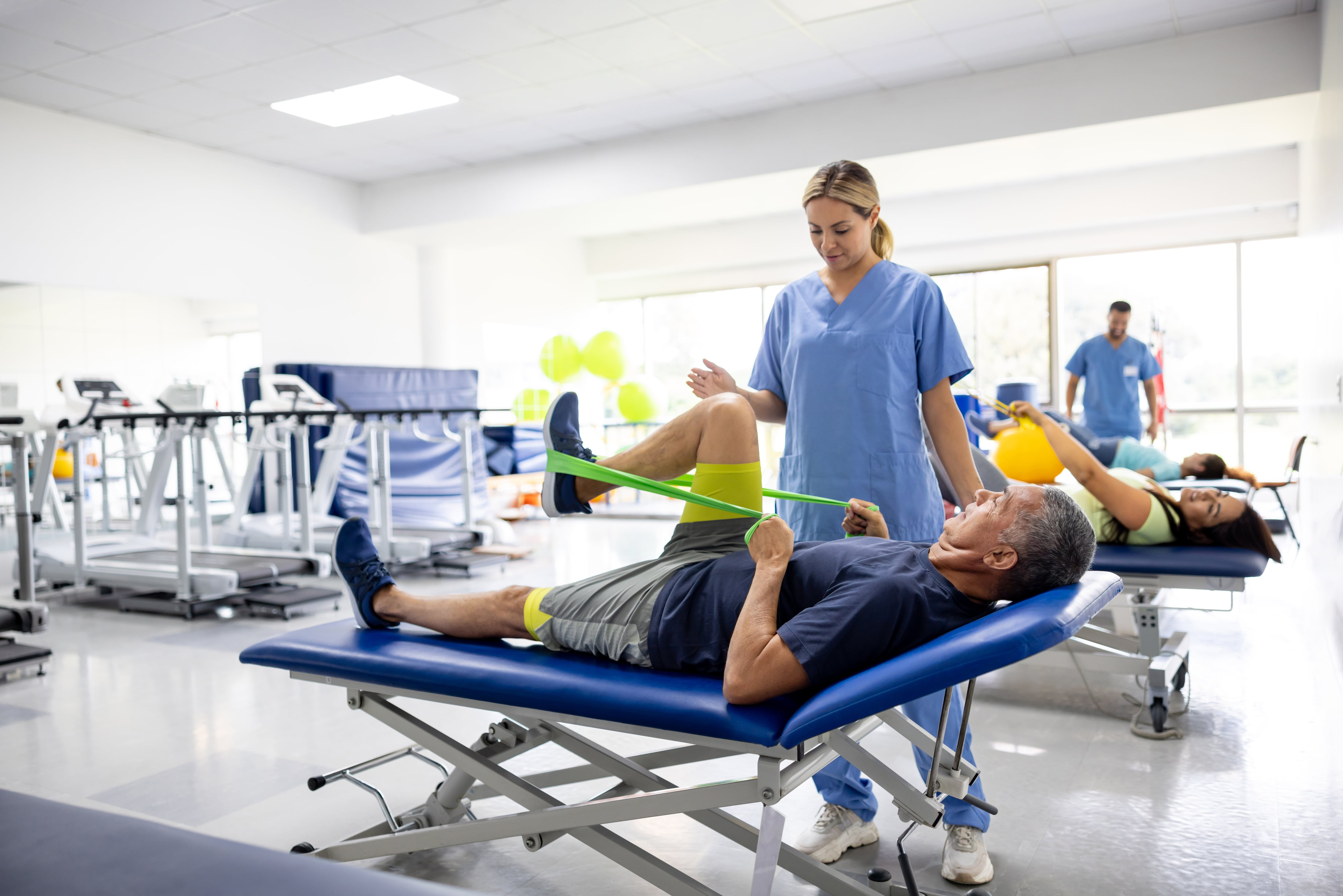 Hombre haciendo ejercicios de terapia física con una banda elástica en compañía de una fisioterapeuta (GettyImages)