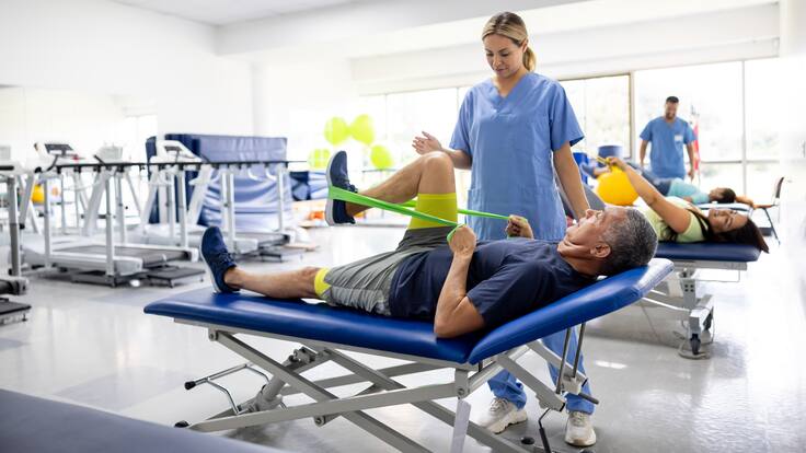 Hombre haciendo ejercicios de terapia física con una banda elástica en compañía de una fisioterapeuta (GettyImages)