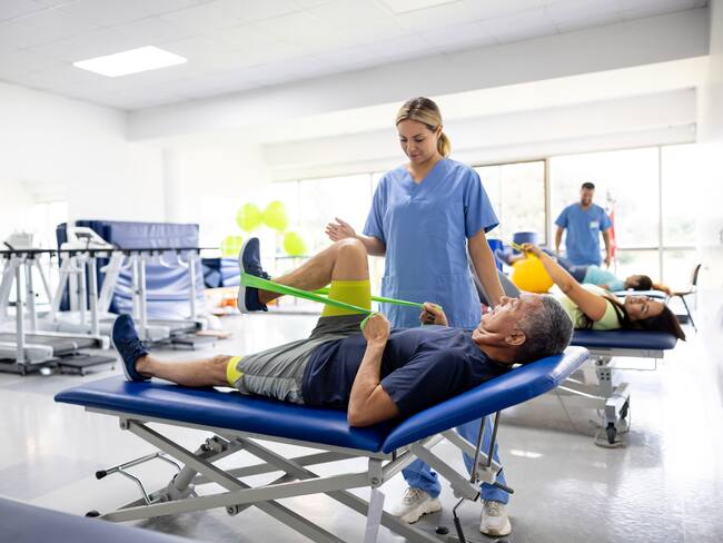 Hombre haciendo ejercicios de terapia física con una banda elástica en compañía de una fisioterapeuta (GettyImages)