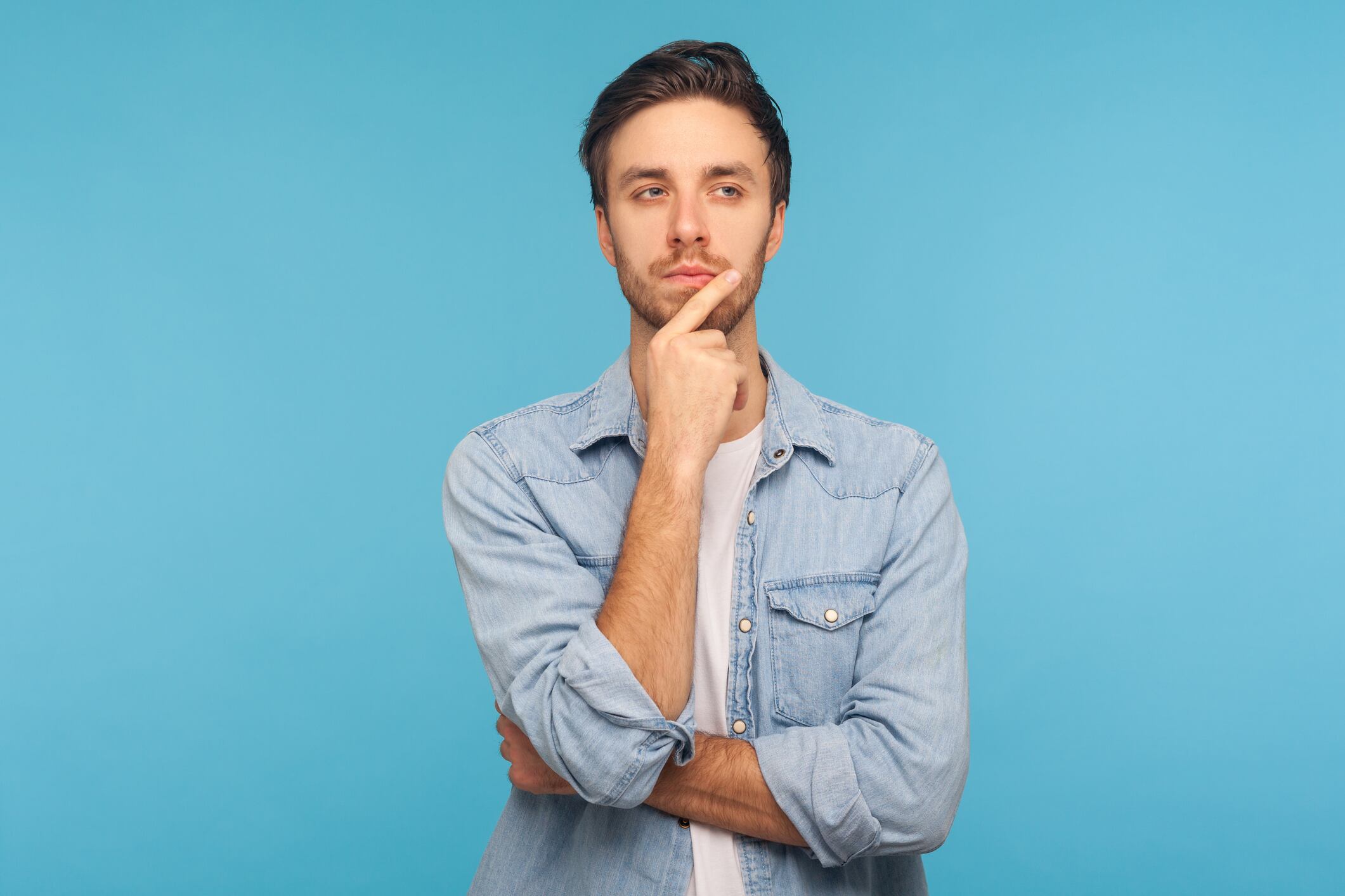 Hmm, let's think! Portrait of pensive handsome man in worker denim shirt touching chin while pondering plan, having doubts about difficult choice, not sure. studio shot isolated on blue background