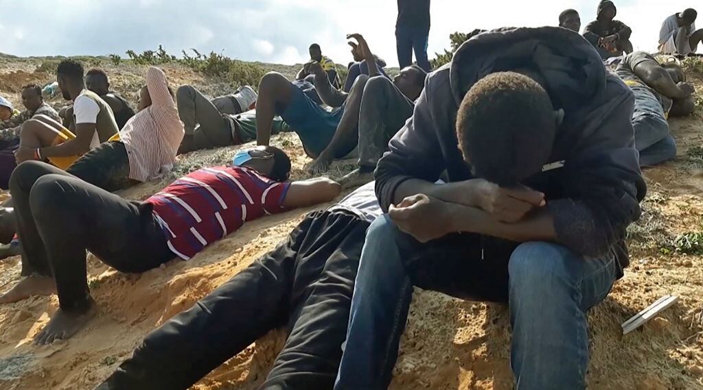 A video grab shows migrant survivors of a deadly shipwreck siting on a sandy beach on the coast of al-Khums, a port city 120 kilometres (75 miles) west of the Libyan capital Tripoli, on November 12, 2020. - According to the United Nations 74 people died in the deadly shipwreck off the Libyan coast, the latest in a spate of migrant vessel sinkings in the central Mediterranean. This year has seen a resurgence of boats in the central Mediterranean, a well-trodden but often deadly route for those hoping to travel to Europe, mainly embarking from Libya and neighbouring Tunisia. (Photo by - / various sources / AFP) (Photo by -/AFP via Getty Images)