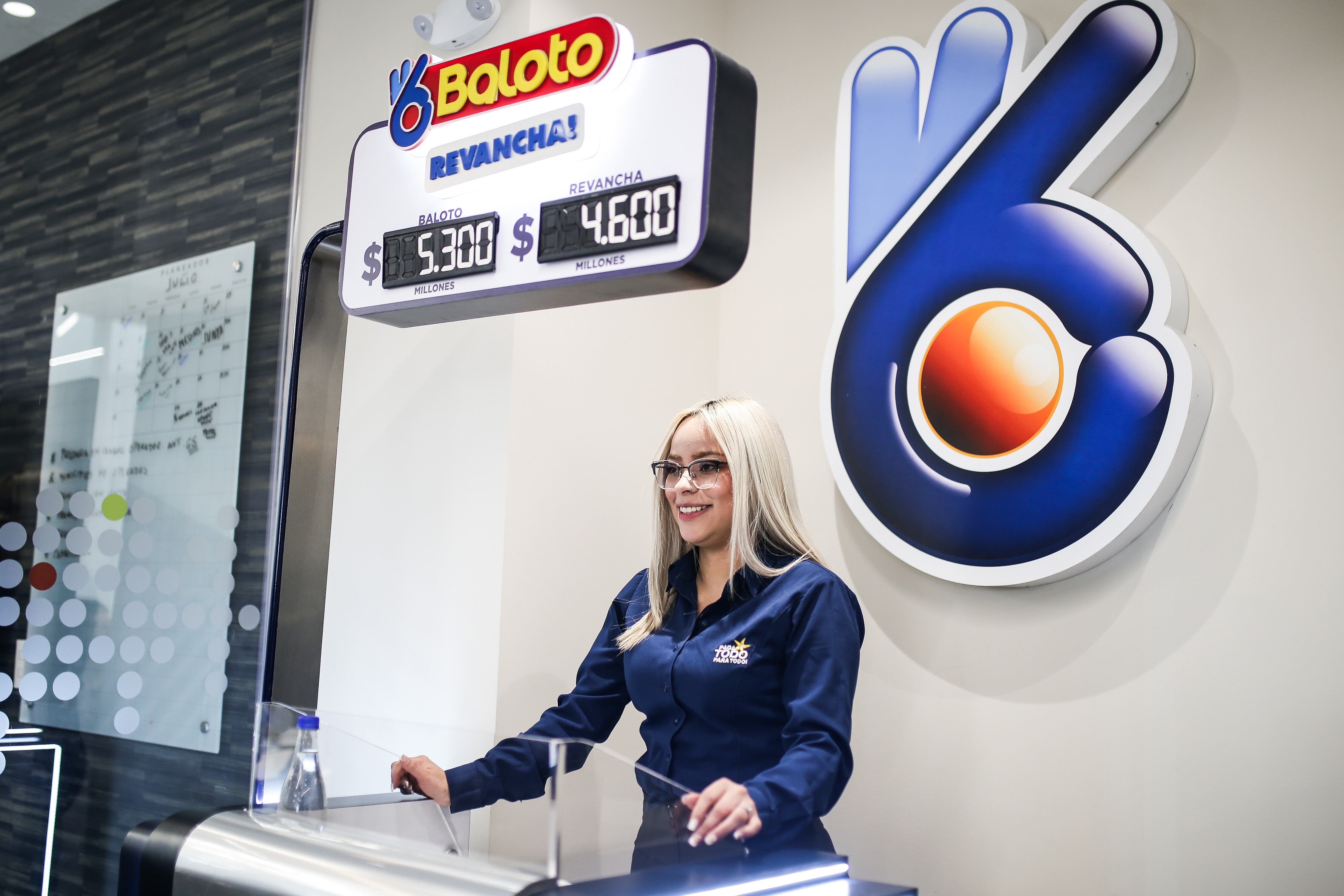 Funcionaria de la entidad Paga Todo y de fondo el tablero con unos valores a jugar de Baloto (Foto vía Colprensa)