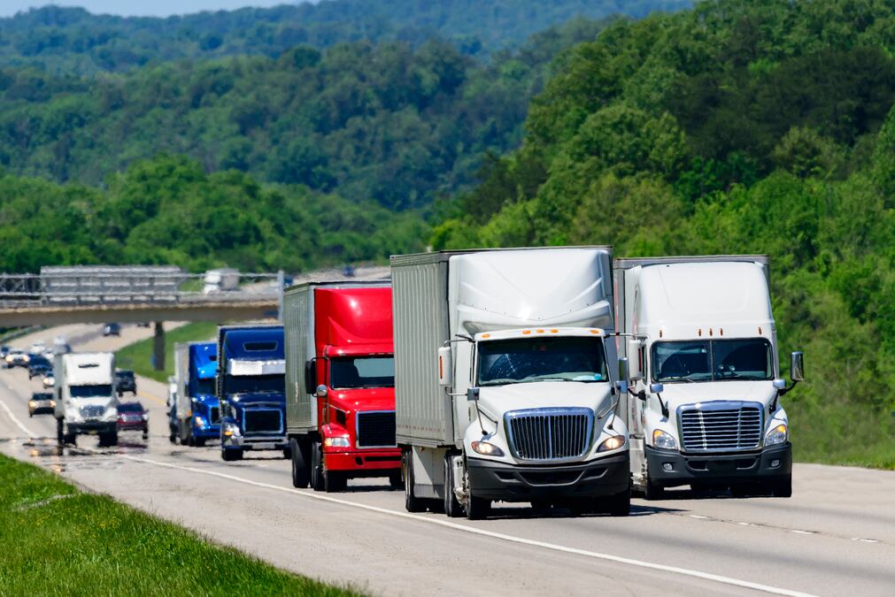 A squadron of eighteen-wheelers lead the way down an interstate highway in eastern Tennessee.  Heat waves rising from the pavement give a shimmering effect to vehicles and forest in the background.
