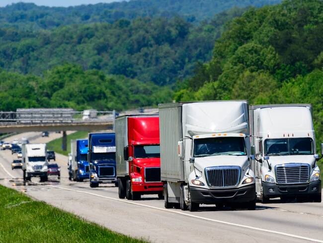 A squadron of eighteen-wheelers lead the way down an interstate highway in eastern Tennessee. Heat waves rising from the pavement give a shimmering effect to vehicles and forest in the background.
