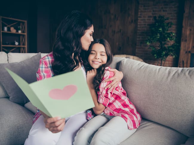 Close up photo two people mum little daughter giving postcard with mommy poem kissing sweet girl unexpected cute delighted wear pink plaid shirts flat apartment room sit cozy couch sofa divan
