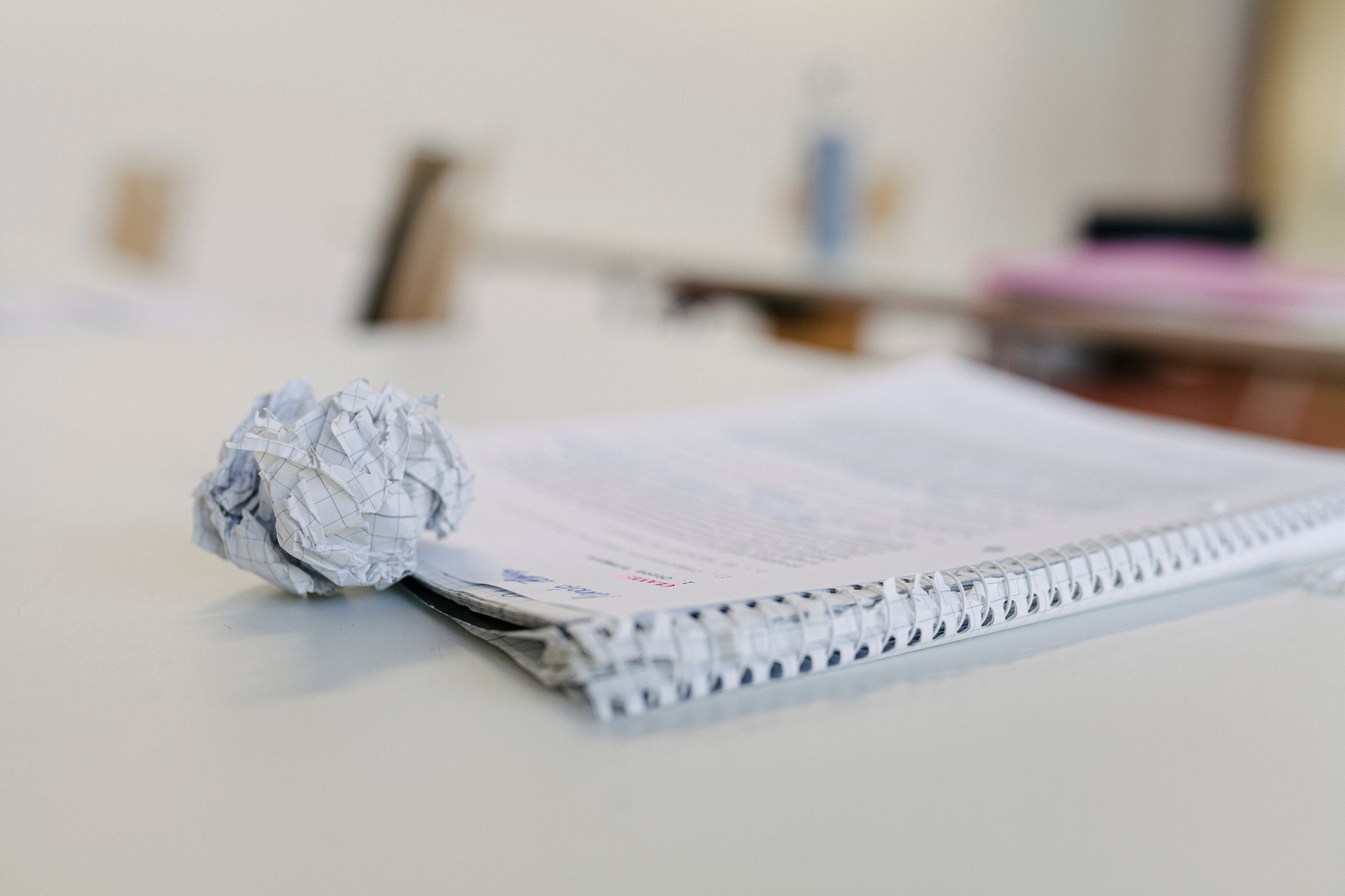 A close up of a scrunched up ball of paper and a students notepad sitting on a high school desk.