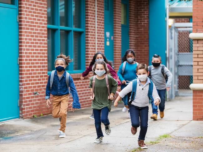 A group of six multi-ethnic elementary school students running outside the school building, carrying backpacks, and wearing face masks. They are back to school during the covid-19 pandemic. The boys and girls are 7 to 10 years old.