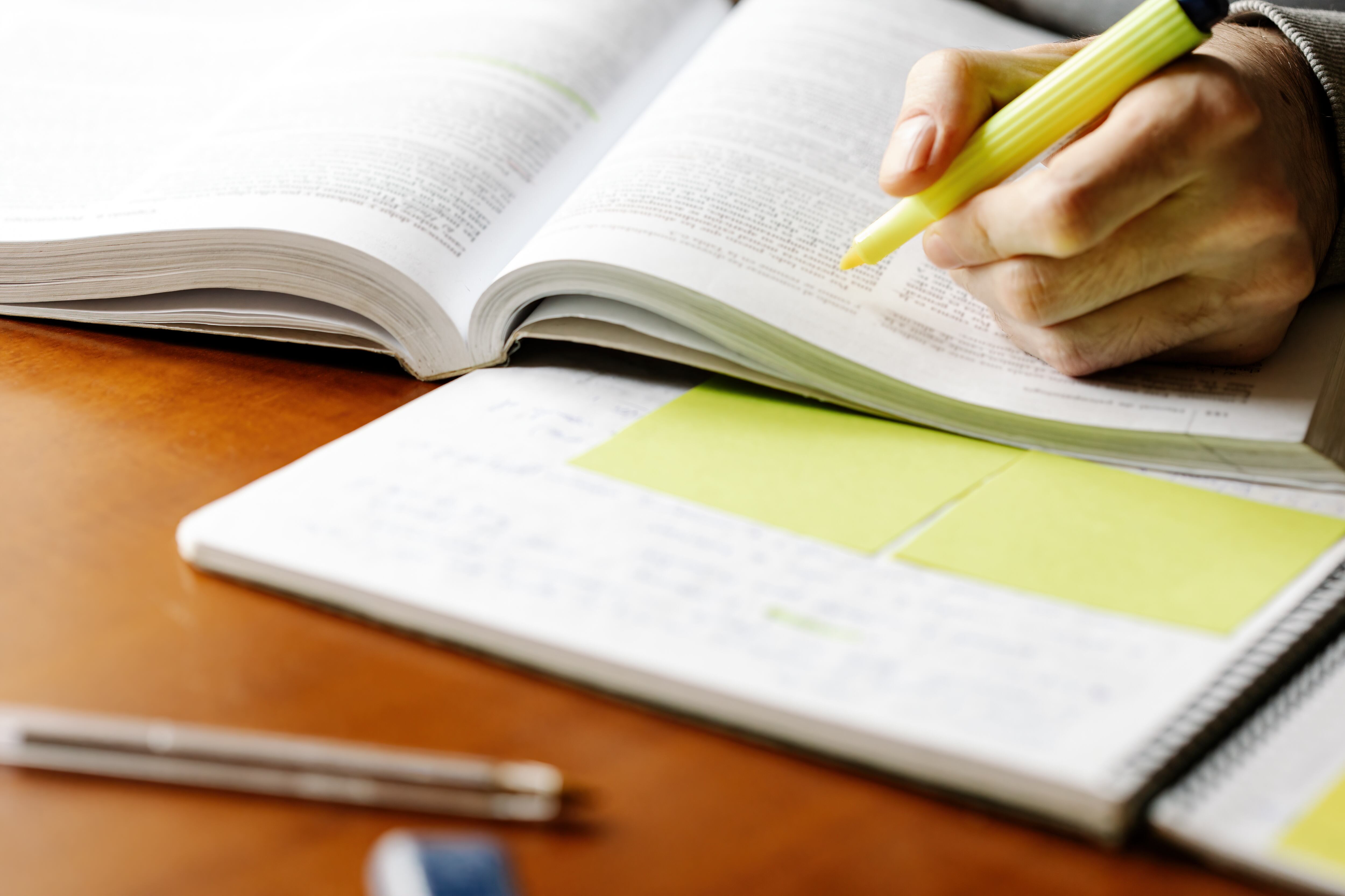 Crop anonymous male teacher with yellow marker pen reading book and making marks while preparing for lesson at table with notebook. Photo: Getty Images