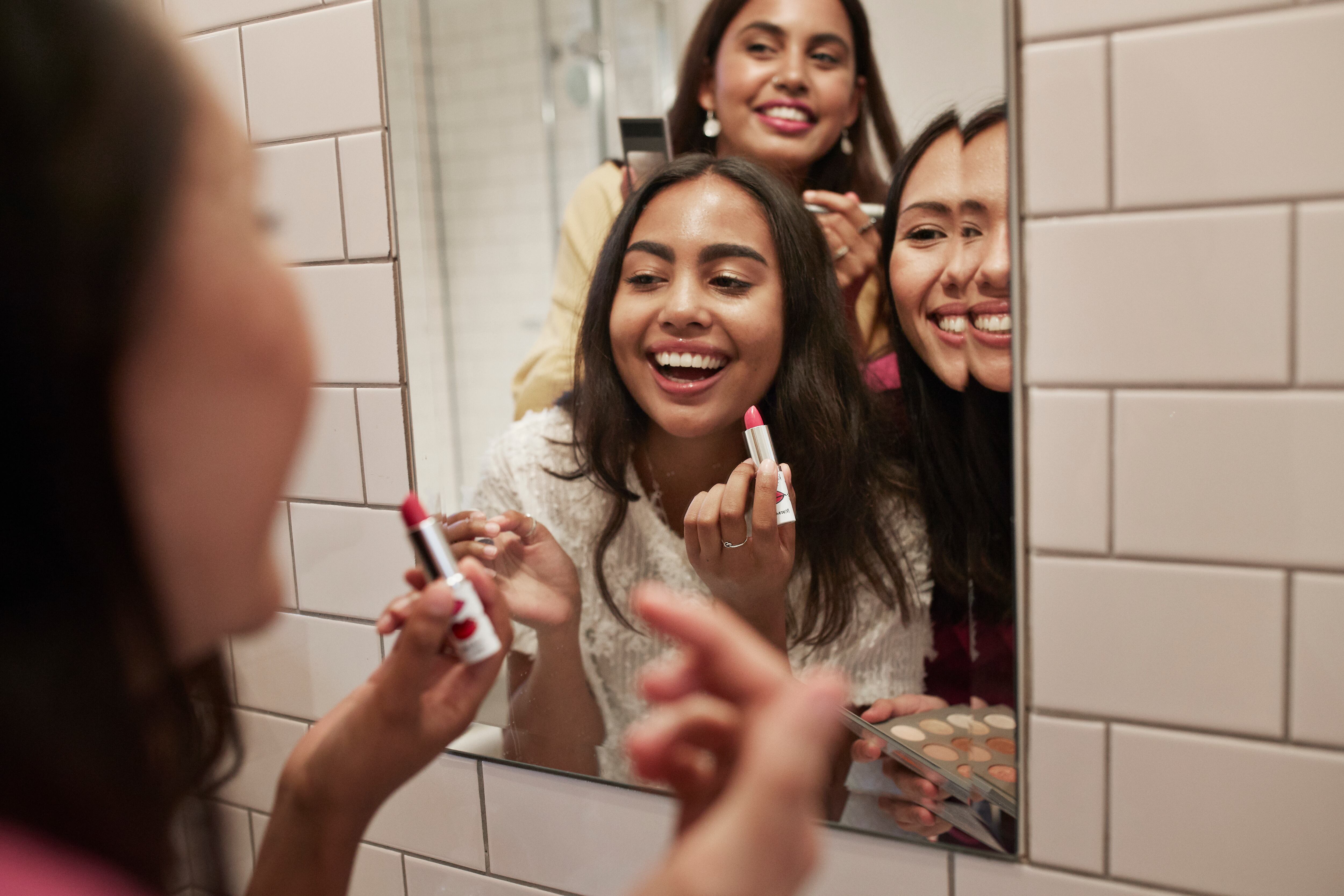 Smiling female friends applying lipstick while reflecting in mirror at home