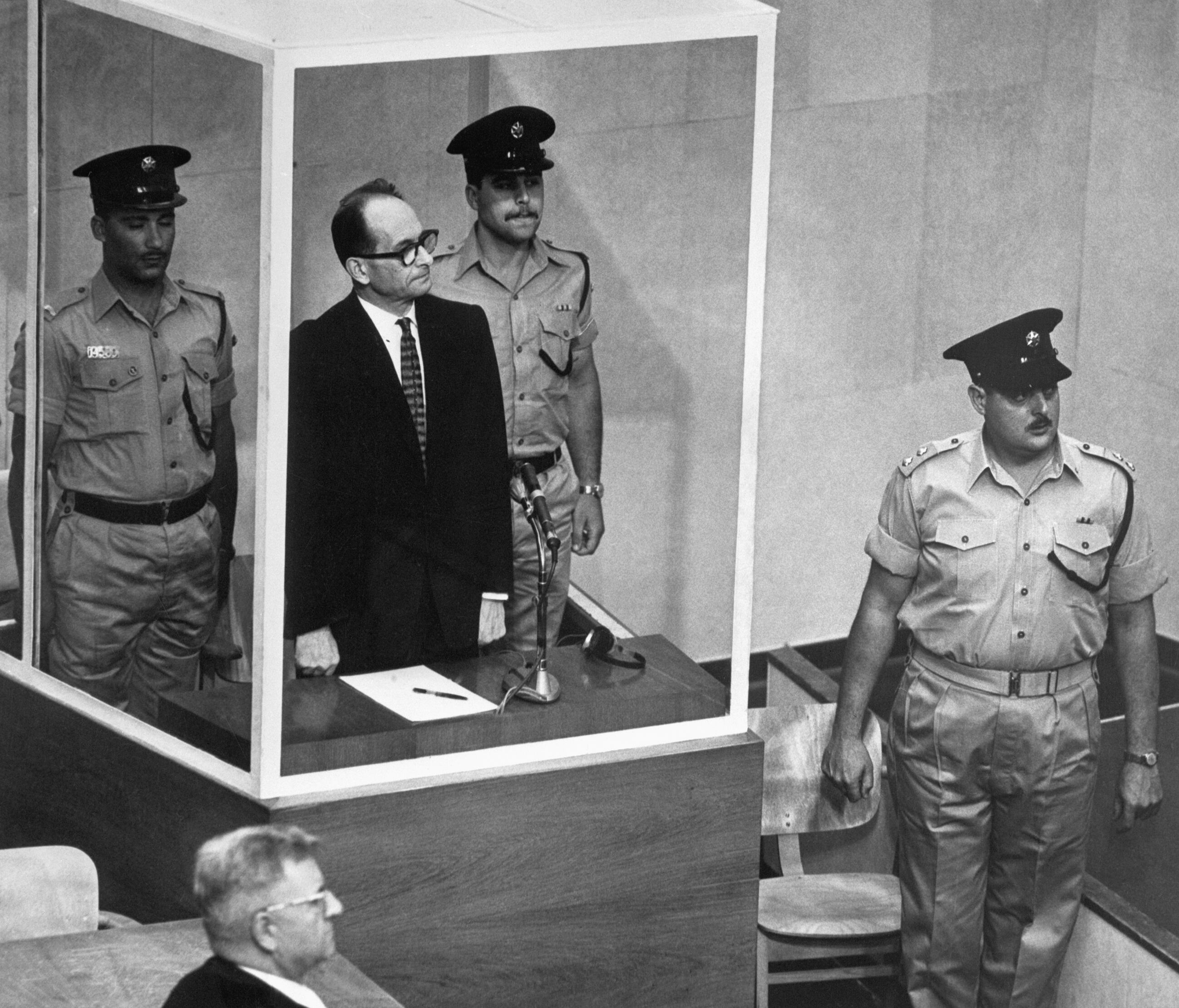 (Original Caption) 5/29/1962-Jerusalem, Israel- Adolf Eichmann, accused Nazi mass murderer, stands in his bullet-proof glass cage to hear Israel's Supreme Court unanimously reject an appeal against his death sentence. With him are two armed guards. The verdict meant that only Israeli President Itzhak Ben-Zvi could save the 56 year old Eichmann, charged with the murder of six million Jews, from the gallows. In the foreground is defense attorney Robert Servatius.