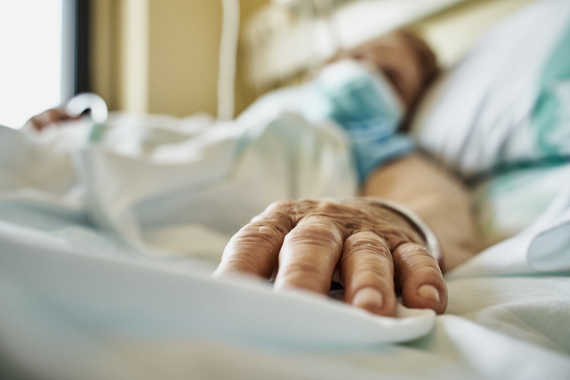 Senior woman wearing mask infected by coronavirus on hospital bed receiving medicine by drip. Close-up fingers of the senior patient ´s hand while she is sleeping. Horizontal photo
