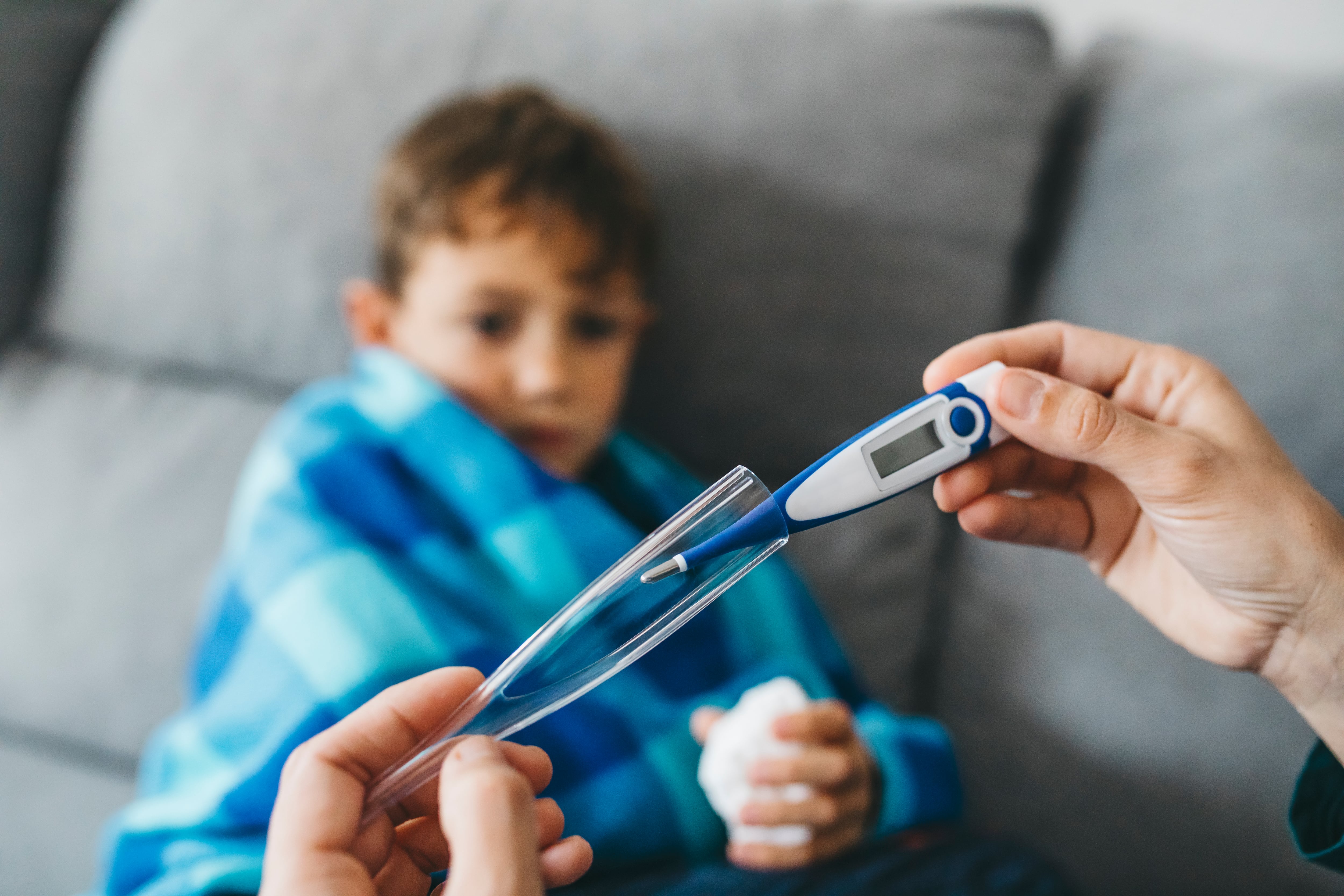 Sick boy sitting on the couch and his mother's hands preparing to place the thermometer in the armpit
