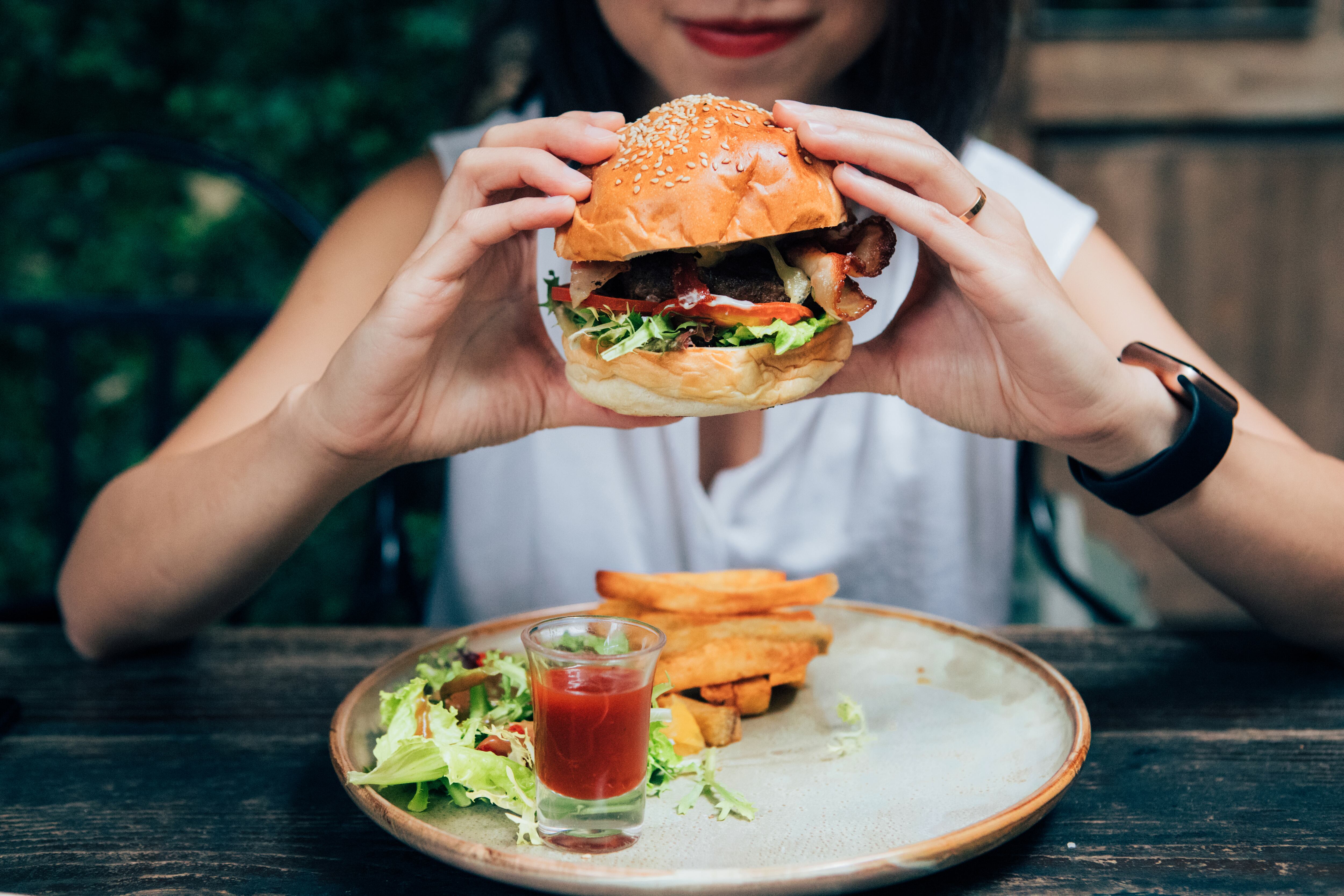 Cropped shot of beautiful young Asian woman eating burger and French fries at the outdoor cafe in the city.