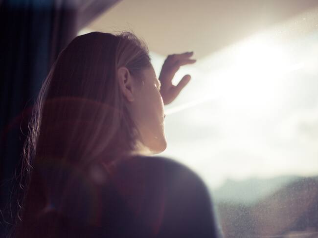 Blondhaired women looking out of window with back light in her face, Spain.