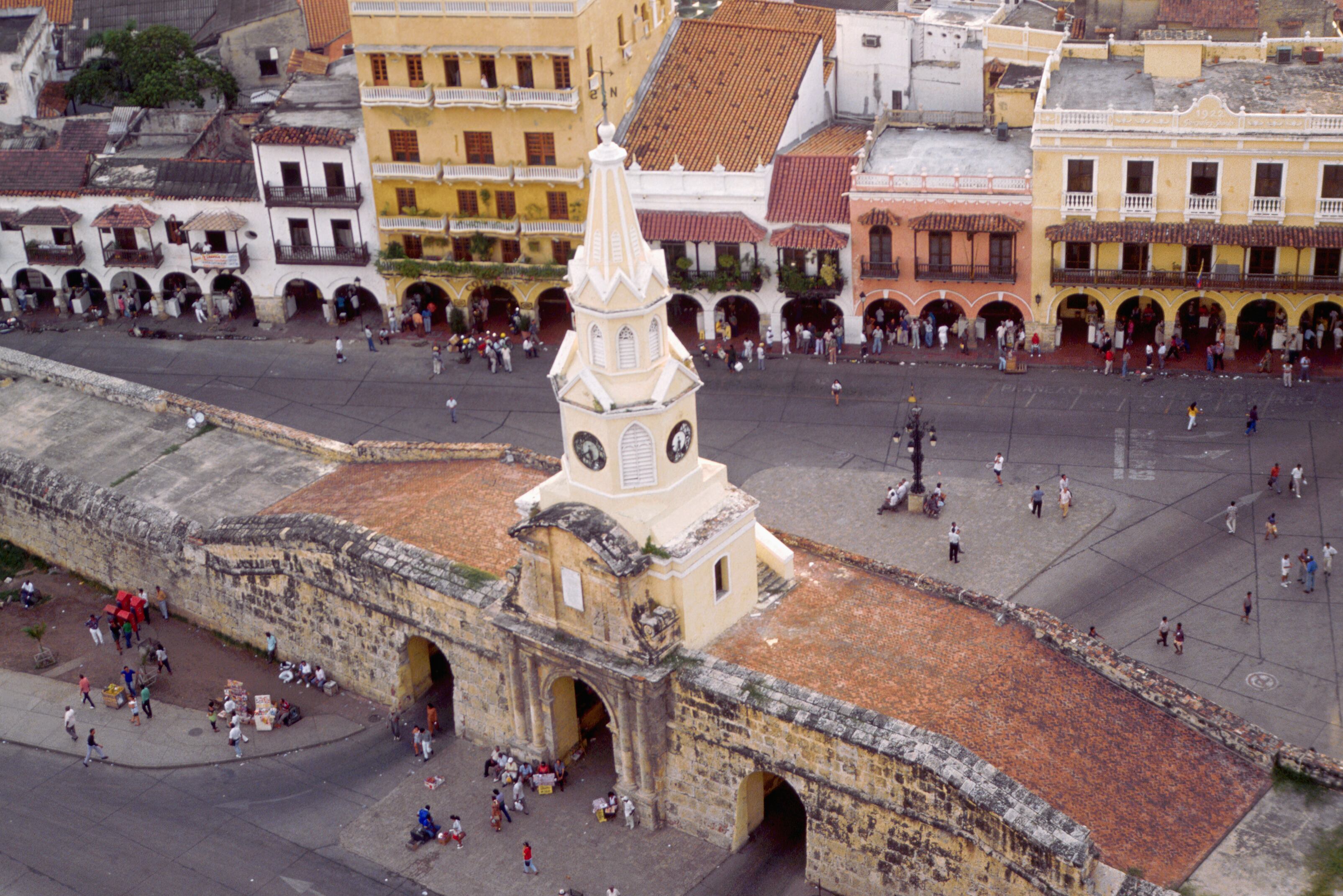 A clock tower positioned on a thick stone wall which cuts through the center of a busy square in the Colombian seaport of Cartagena.