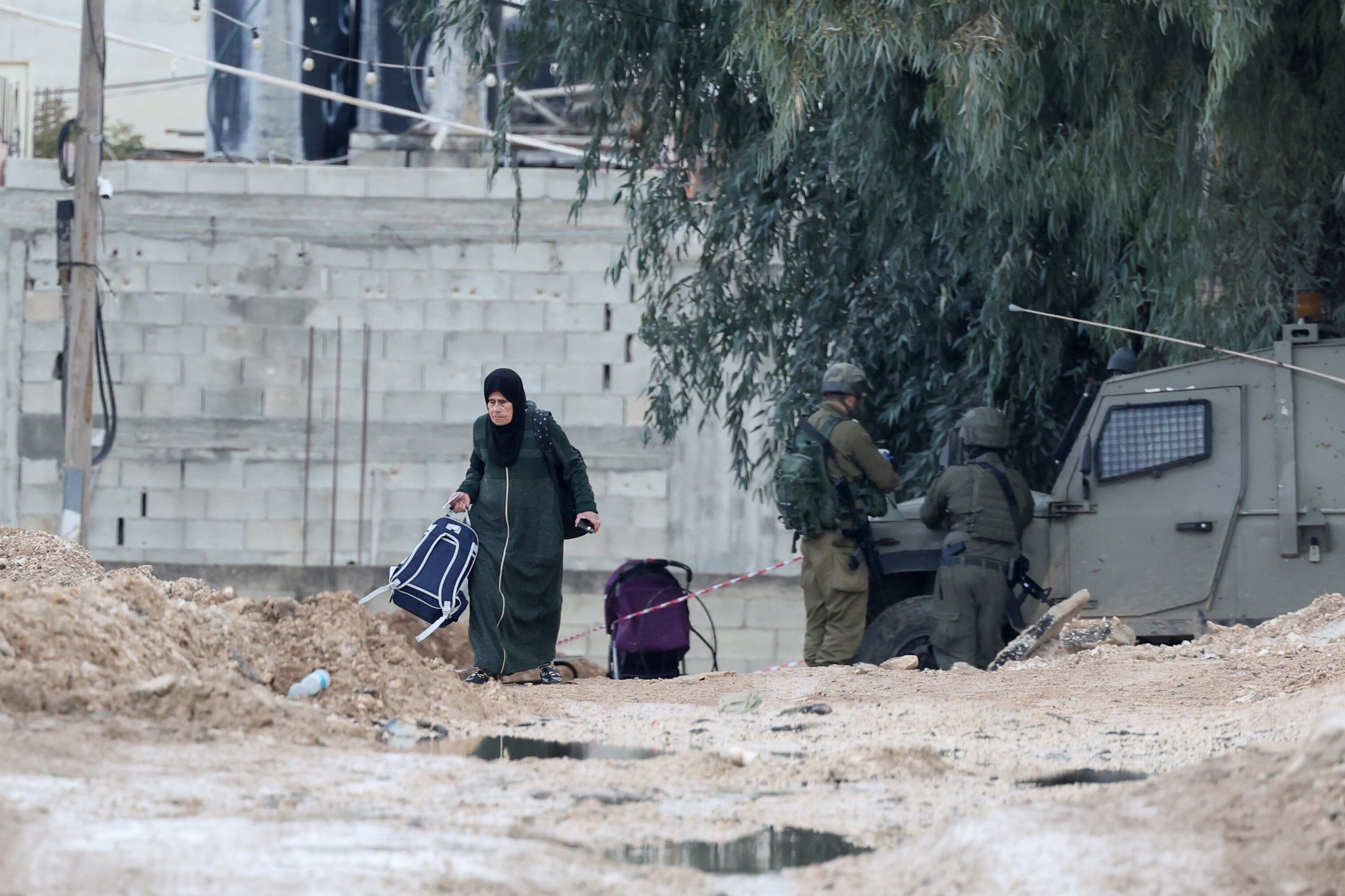 Yenín (-), 23/01/2025.- Una mujer palestina con sus pertenencias pasa junto a soldados israelíes al salir del campo de refugiados de Yenín en el tercer día de una operación militar israelí.