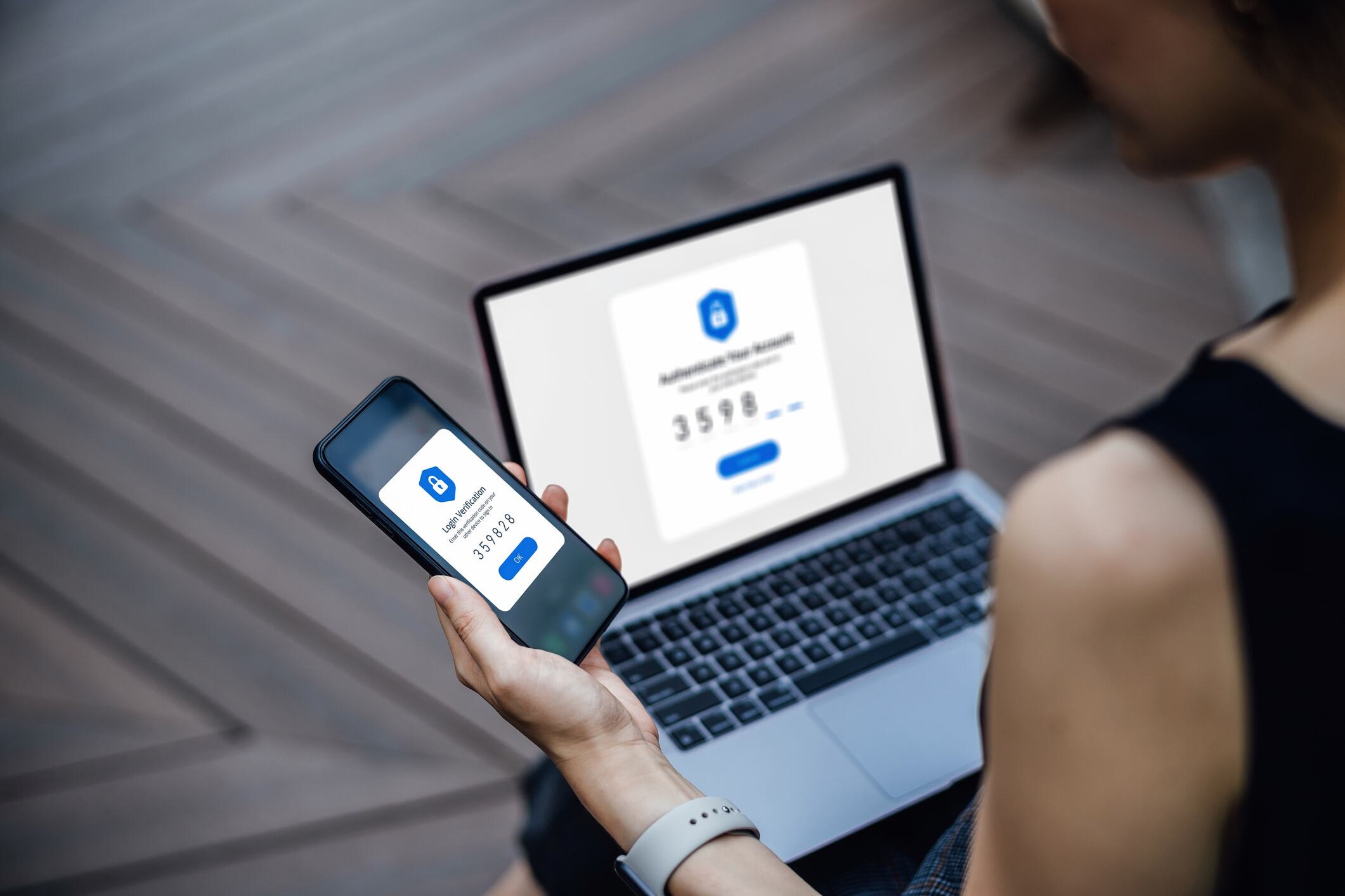 Over the shoulder view of young woman using mobile device with Two-Factor Authentication (2FA) security while logging in securely to her laptop. Privacy protection, internet and mobile security
