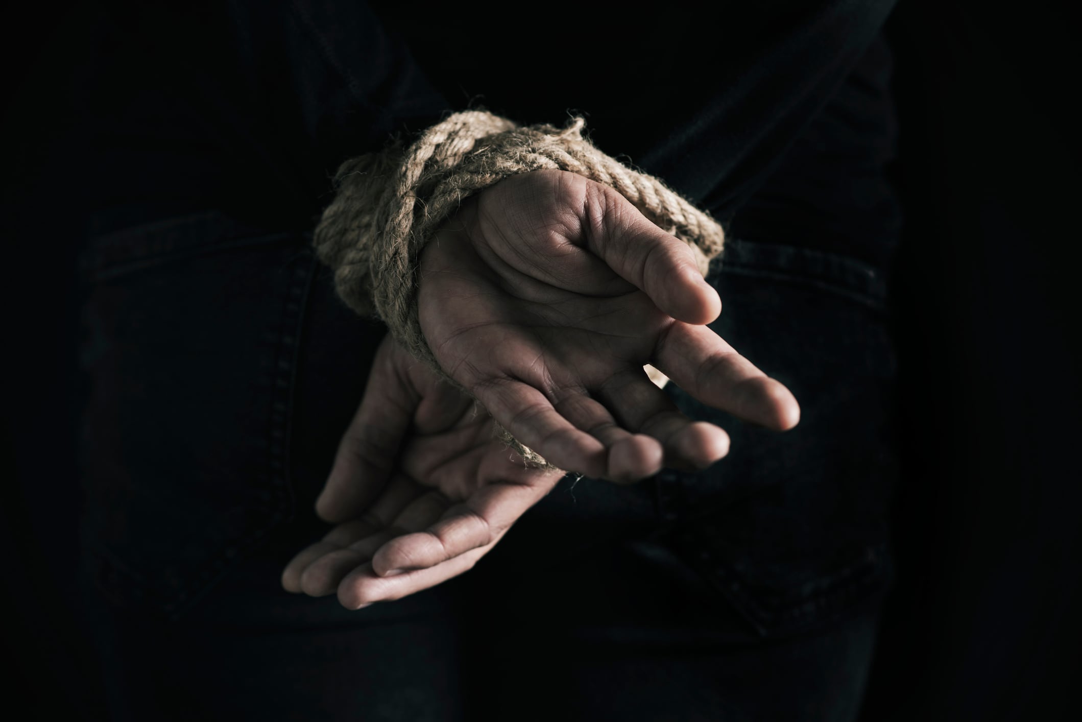closeup rear view of a man with his hands tied behind his back with rope, against a black background