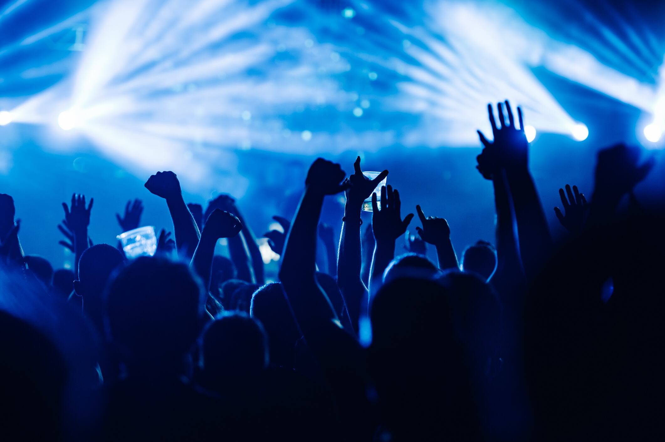 Silhouettes of people rising their hands and dancing on an open air music festival. There are blue stage lights. People having fun at rock concert on a summer music festival.