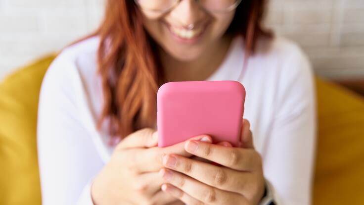 Mujer de camisa blanca utilizando aplicaciones en su celular mientras sonríe (Foto vía Getty Images)