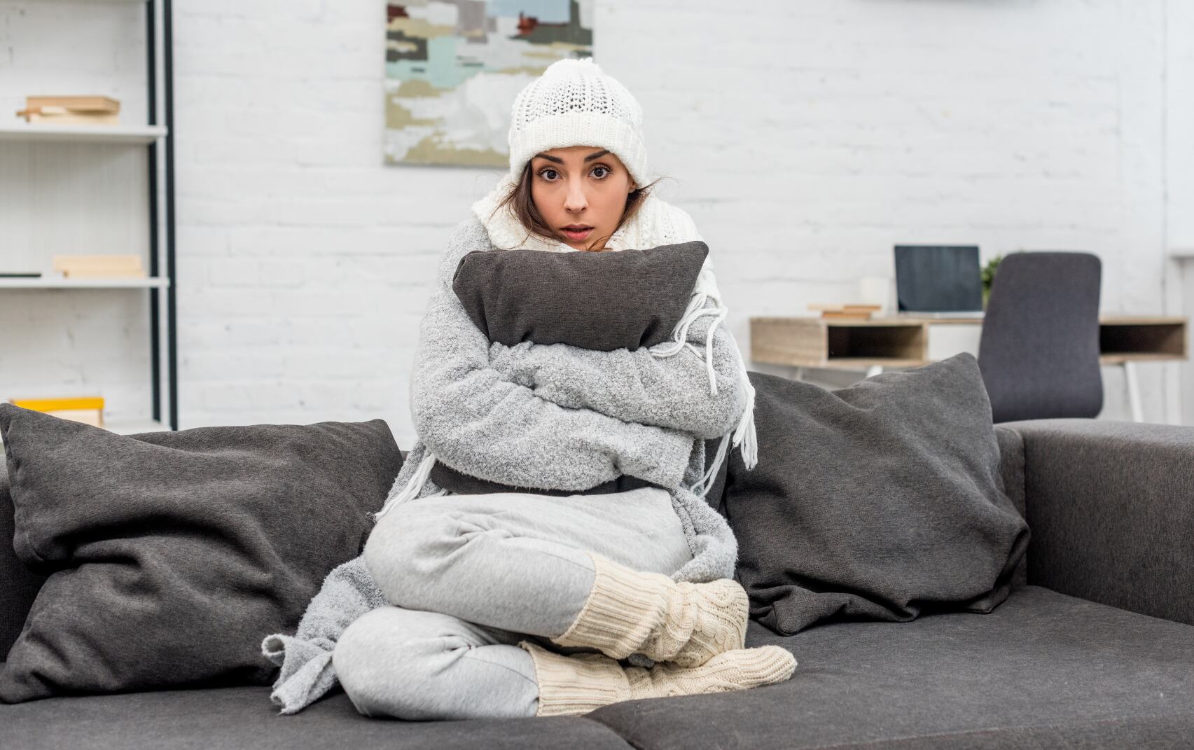 Mujer con frío, vistiendo gorro abrigo y abrazando un cojín debido a las najas temperaturas en su apartamento (Foto vía Getty Images)