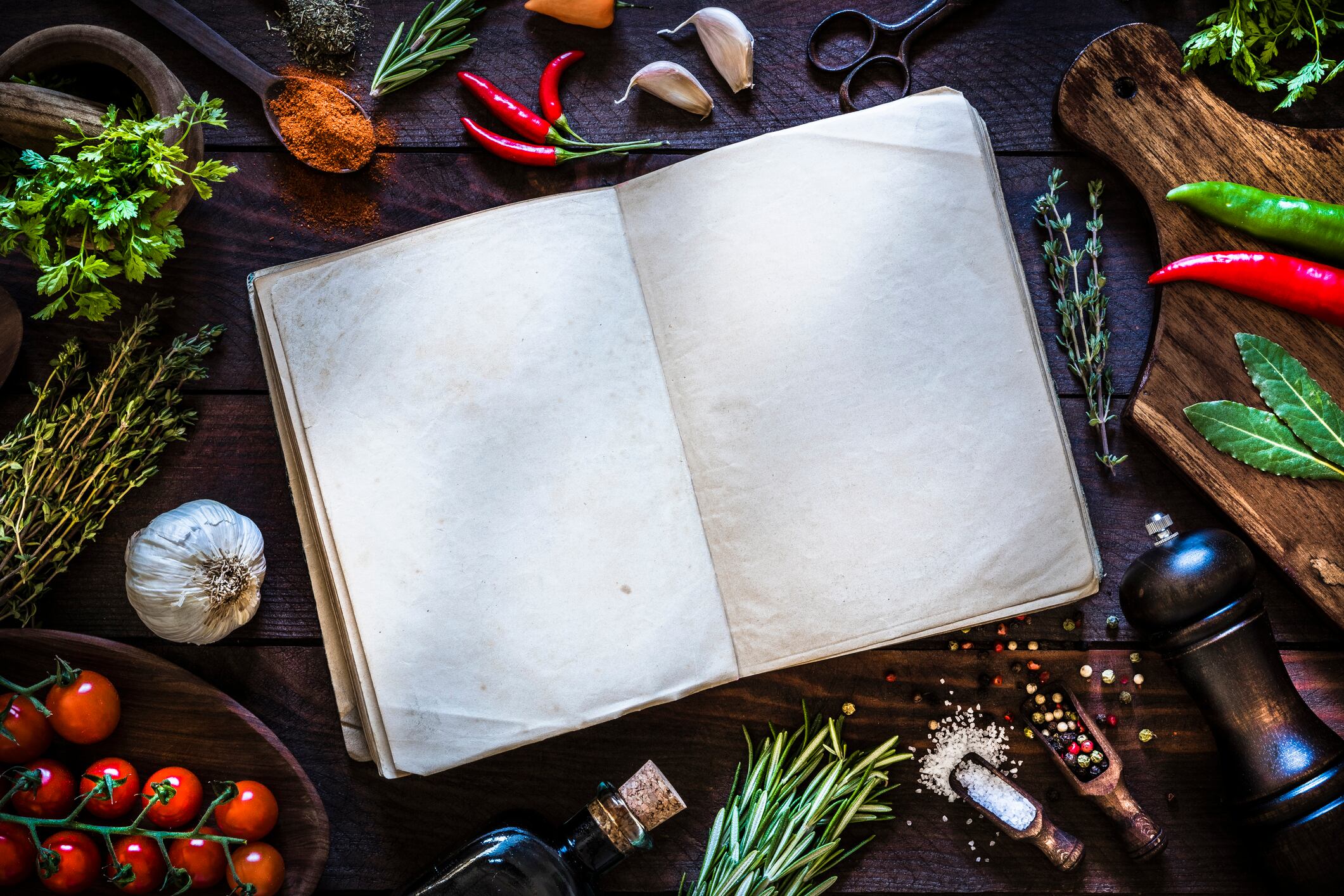 Top view of a vintage open cookbook surrounded by some spices and herbs like thyme, rosemary, laurel, pepper, garlic and chervil, and some kitchen utensils. The cookbook is at the center of the image and its pages are empty so you can use it as a useful copy space. Objects are on a rustic dark brown wooden table. Low key DSLR photo taken with Canon EOS 6D Mark II and Canon EF 24-105 mm f/4