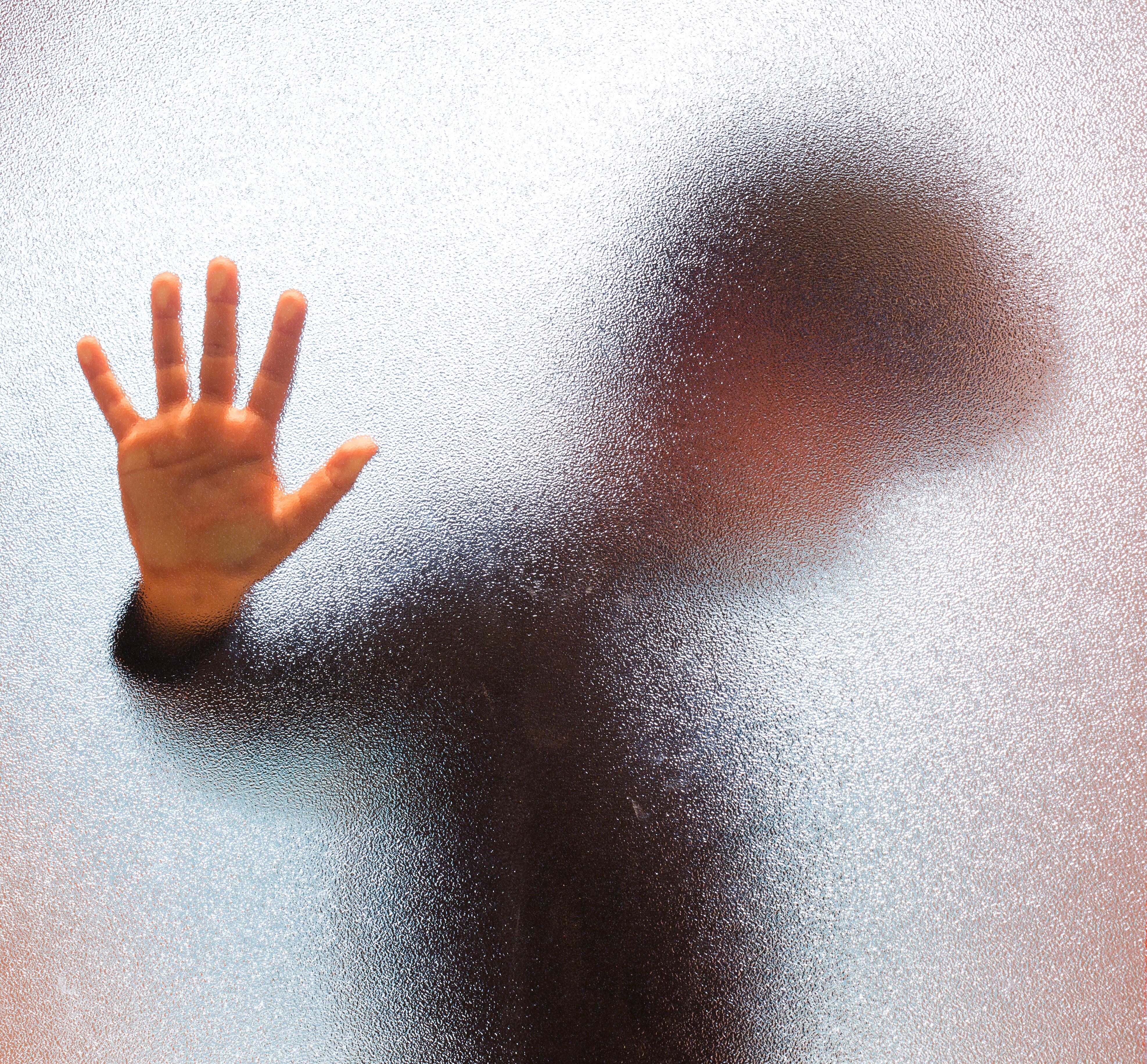 Blurred sad boy leaning open hand against glass door.