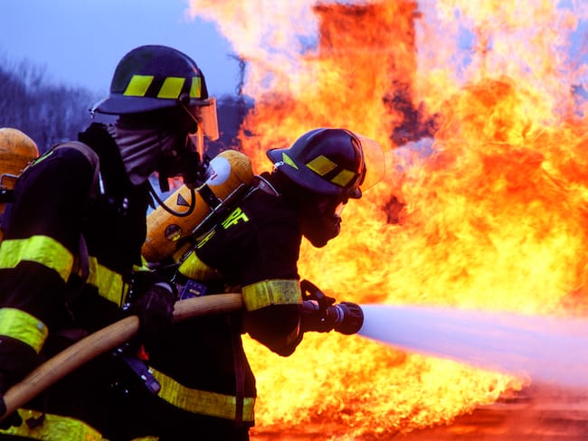 Two fireman spraying water at base of orange flames.