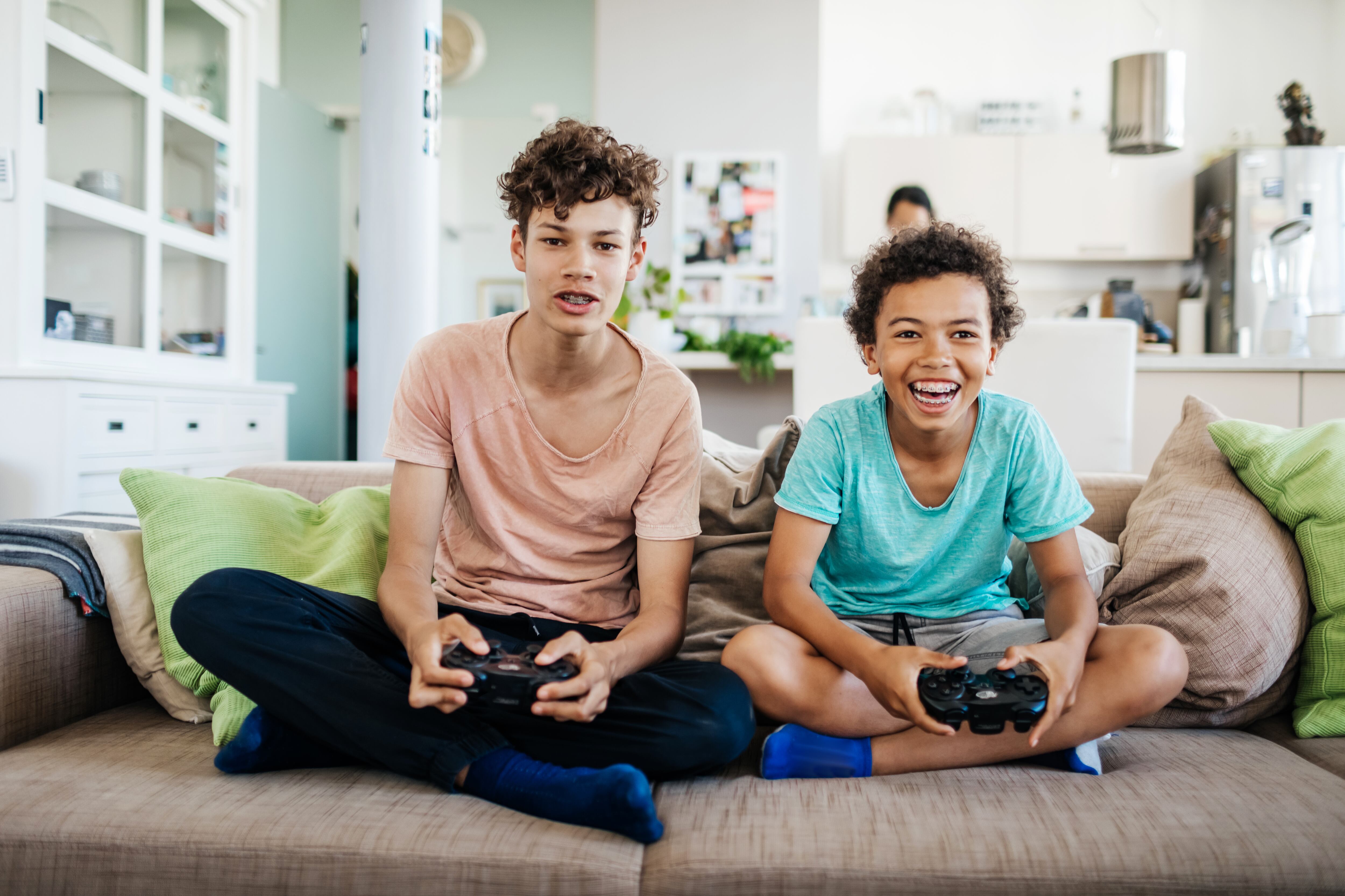Two young brothers sitting on the couch at home, smiling while playing computer games together.