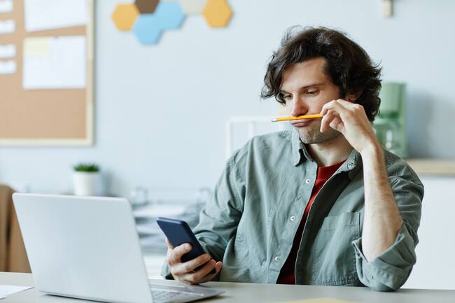 Hombre adulto, mirando el celular mientras, juega con un lápiz en el trabajo. (Getty Images)